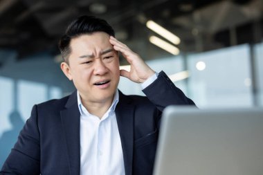 Ciddi genç Asyalı adam masanın başında oturuyor, kafasını tutuyor ve dizüstü bilgisayarına endişeli bir şekilde bakıyor..