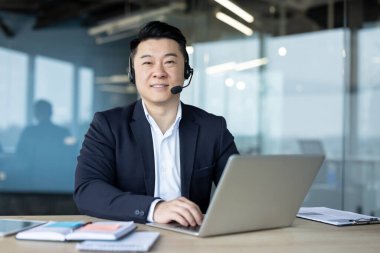 Ofiste dizüstü bilgisayarla çalışan ve kameraya bakan takım elbiseli, gülümseyen Asyalı genç bir adamın portresi..