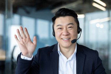 Takım elbiseli ve kulaklıklı gülümseyen Asyalı genç bir adamın yakından çekilmiş bir portresi. Kameraya bakıyor, selamlıyor ve el sallıyor..