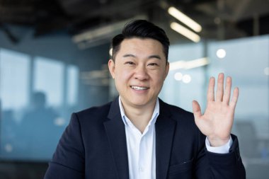 Ofiste, kameranın önünde takım elbise giymiş gülümseyen ve elini sallayan Asyalı genç bir adamın yakın plan portresi..