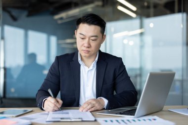 Ciddi ve odaklanmış genç Asyalı bir adam ofiste elinde dizüstü bilgisayarla oturuyor ve belgelerle çalışıyor..