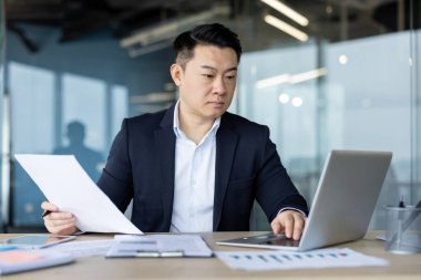 Ciddi bir Asyalı genç işadamı ofiste masa başında oturuyor ve bir dizüstü bilgisayar üzerinde çalışıyor, elinde belgeler ve bir klavye üzerinde daktilo kullanıyor..