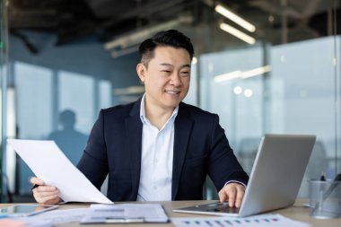 Gülümseyen Asyalı genç adam, ofis çalışanı takım elbise içinde iş yerinde oturuyor, dizüstü bilgisayarla ve belgelerle çalışıyor..