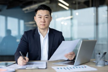 Ofis ortamında çalışan Asyalı bir işadamı. O bir takım elbise giyiyor ve masasında belgeler üzerinde çalışıyor. Ona odaklan..