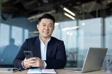 Genç, gülümseyen ve başarılı bir Asyalı iş adamının portresi. Ofiste dizüstü bilgisayarla oturuyor, elinde telefon, kameraya bakıyor..