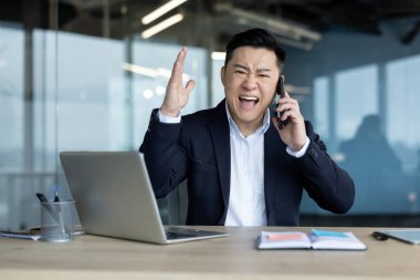 Kızgın genç Asyalı bir adam ofiste masada oturuyor, telefonda konuşuyor ve bağırıyor..