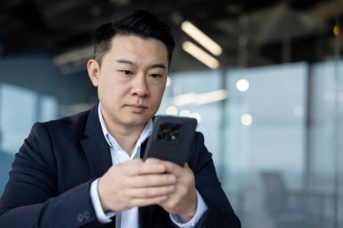 Ofiste oturup cep telefonunu kullanan genç, odaklanmış ve ciddi bir Asyalı iş adamının yakın plan fotoğrafı..