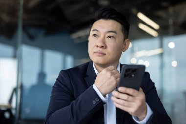 İş yerinde takım elbiseli genç bir Asyalı 'nın yakın plan fotoğrafı. Elinde bir telefonla oturmuş, ciddi ciddi camdan dışarı bakıyor..