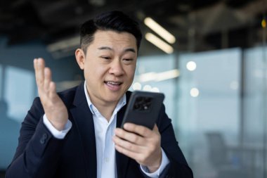 Takım elbiseli genç bir Asyalının yakın plan fotoğrafı. Masada oturuyor ve telefonla konuşuyor..