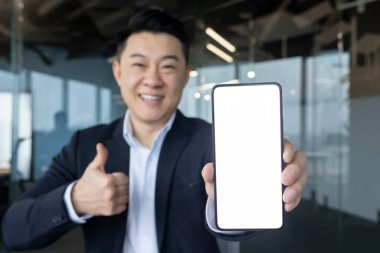 Takım elbiseli gülümseyen Asyalı genç bir adamın portresi kameraya cep telefonunun beyaz ekranını gösteriyor ve kameraya parmağıyla süper bir jest yapıyor..