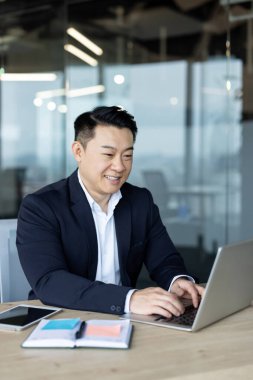 Ofiste dizüstü bilgisayarla çalışan, gülümseyen ve başarılı Asyalı iş adamının dikey fotoğrafı..