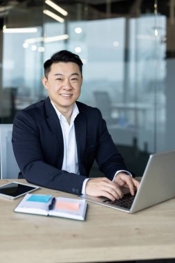 Gülümseyen ve başarılı Asyalı genç bir iş adamının dikey fotoğrafı. Ofiste oturmuş dizüstü bilgisayarla çalışıyor..