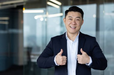Ofiste takım elbiseli gülümseyen Asyalı genç bir adamın portresi ve parmaklarıyla kameraya doğru süper bir jest yapıyor..