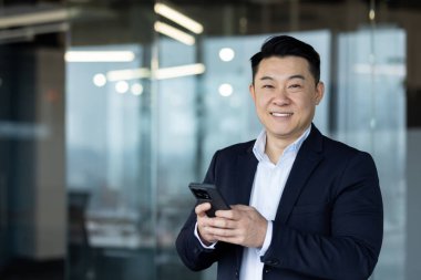 Genç, gülümseyen Asyalı bir işadamının portresi, bir firma ve şirketin kurucusu, ofiste duruyor, bir telefon tutuyor ve kameraya bakıyor..