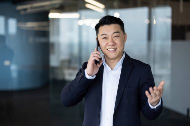 Gülümseyen Asyalı genç işadamı ofiste cep telefonuyla konuşuyor ve kameraya bakıyor..