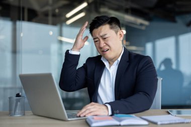 Frustrated and upset young asian man sitting in office at desk and working on laptop gesturing with hand.
