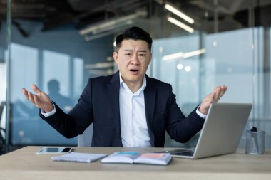 Portrait of an upset young Asian man in a suit sitting at the desk in the office and waving his arms, at the camera in frustration.