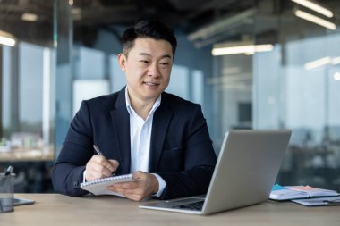A young Asian man in a suit is sitting at a table, looking at the laptop screen and making notes in a notebook.