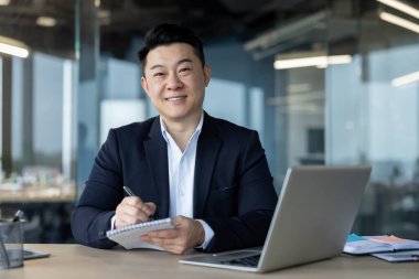 Portrait of a young Asian man working in an office, sitting at a desk, working on a laptop and taking notes in a notebook, looking at the camera.