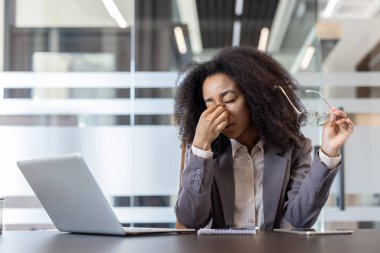 Tired and overwhelmed young African American businesswoman sitting at a desk in the office, holding glasses in her hand and rubbing her eyes with her hand.