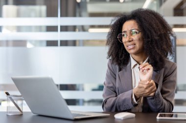African American young woman working in an office, sitting at a desk, grimacing from boredom, holding her wrist with her hand.