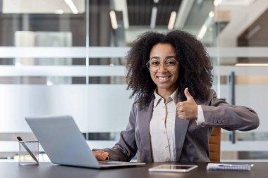 Portrait of a smiling young African American woman in a suit sitting at a desk in the office, looking at the camera and showing a super gesture with her finger.