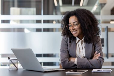 Photo of a smiling young African American businesswoman in a suit sitting at an office desk with her arms crossed, and looking at the laptop screen.