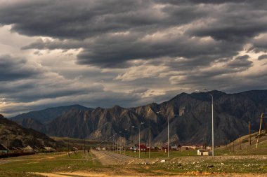 Kara fırtına bulutları ile bir dağ manzara fonuna karşı küçük bir yerleşim ve yeşil çim geçen Asfalt yol.