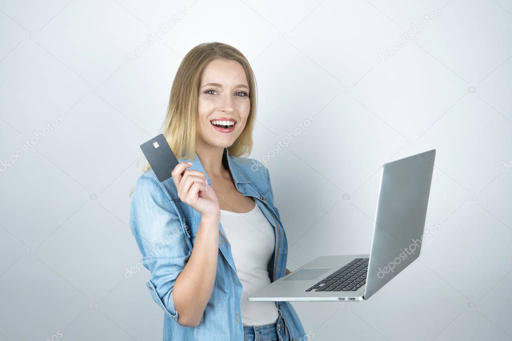 beautiful blond woman looks happy holding her bank card in one hand and laptop in another , online shopping, isolated white background