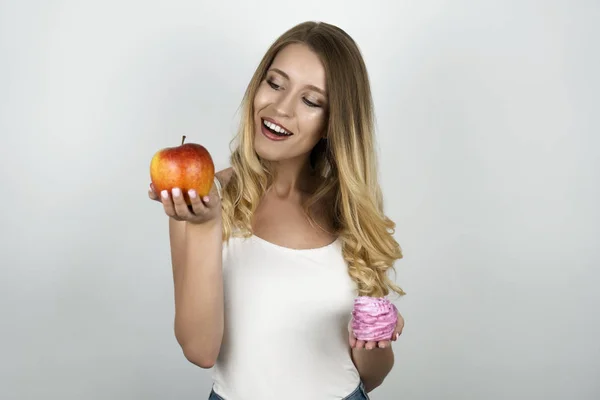 Leende blond attraktiv kvinna som håller saftigt rött äpple i ena handen och rosa välsmakande cupcake i andra hand isolerad vit bakgrund — Stockfoto