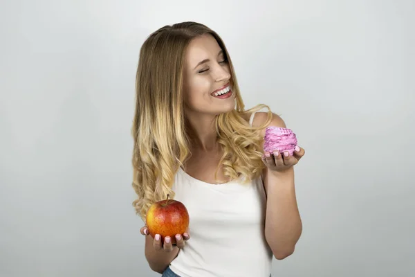 Ung blond attraktiv kvinna som håller saftigt rött äpple i ena handen och rosa välsmakande cupcake i andra hand isolerad vit bakgrund — Stockfoto