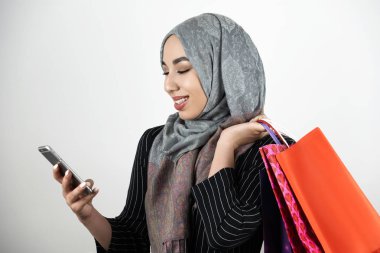 genç güzel gülümseyen Müslüman iş kadını türbantürban türban bir eliyle akıllı telefon dokunarak ve başka bir elinde izole beyaz arka plan alışveriş çantaları taşıyan giyen
