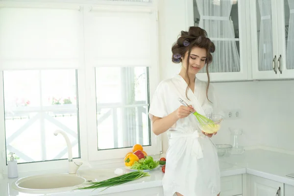 Vacker ung brunett i hår papiljotter bär vit dressing klänning blandning ägg förbereder frukost på morgonen i hennes ljusa kök — Stockfoto
