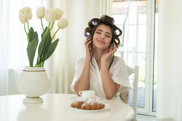 Hermosa joven morena sonriente en rulos de pelo escuchando música mientras desayuna en cocina luminosa —  Fotos de Stock