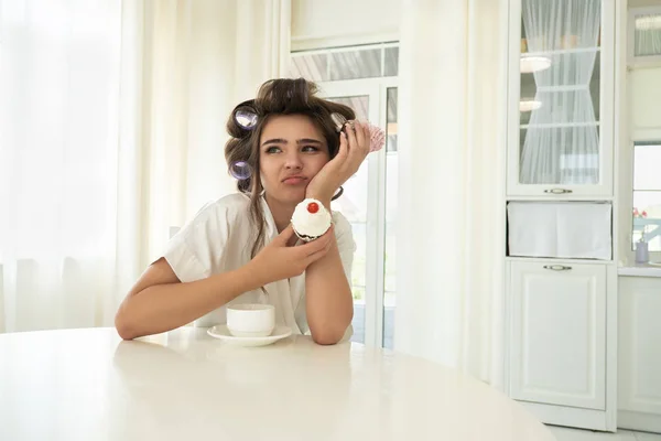 Bella giovane donna bruna in bigodini che tengono i cupcake in entrambe le mani seduti con una tazza di caffè in cucina luminosa cercando sconvolto — Foto Stock