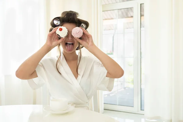 Hermosa joven morena en rizadores de pelo mostrando la lengua sosteniendo dos cupcakes en ambas manos cerca de sus ojos sentado con taza de café en la cocina brillante —  Fotos de Stock