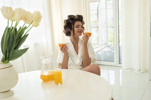 Belle jeune femme brune dans les bigoudis de cheveux tenant des oranges dans les deux mains dans la cuisine lumineuse — Photo