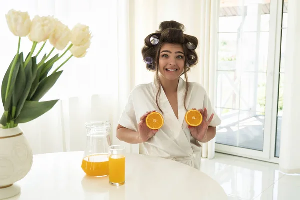 Belle jeune femme brune dans les bigoudis de cheveux tenant des oranges dans les deux mains près de ses seins dans la cuisine lumineuse regardant surpris — Photo