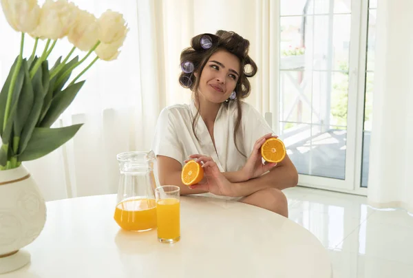 Bella giovane donna bruna sorridente in bigodini tenendo le braccia con le arance incrociate seduta in cucina luminosa — Foto Stock