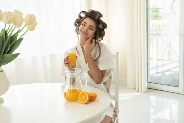 Atractiva joven morena mujer en rulos de pelo disfrutando de naranja fresco exprimido jugo sentado en la cocina brillante teniendo desayuno saludable —  Fotos de Stock