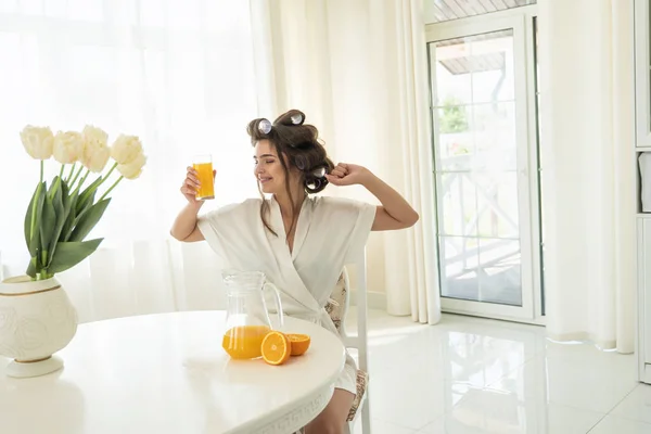 Atractiva joven morena mujer en rulos de pelo disfrutando de naranja fresco exprimido jugo estiramiento después de dormir en la cocina brillante —  Fotos de Stock