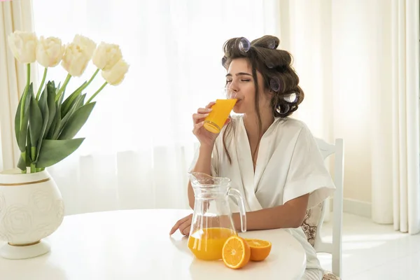 Bella giovane donna bruna in bigodini di capelli godendo arancia succo spremuto fresco bevendolo da vetro seduto in cucina luminosa — Foto Stock