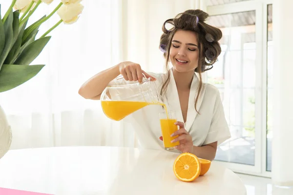 Vacker ung brunett kvinna i hår papiljotter hälla apelsin färskpressad saft från tillbringare i glas sittande i ljust kök — Stockfoto