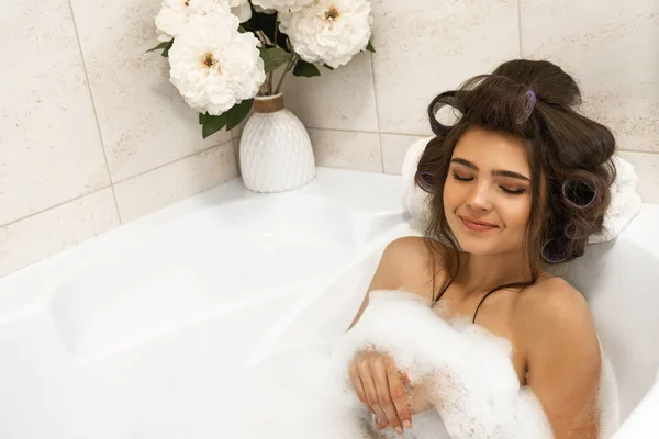 Atraktivní mladá uvolněná brunetka ve vlasech, která si vychutnávala koupel s pěnou v zářivě koupelně — Stock fotografie