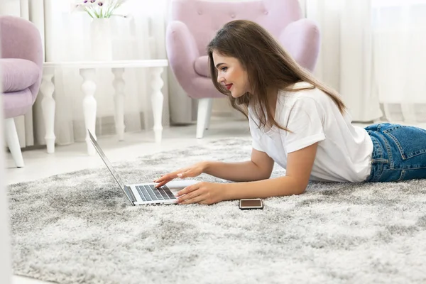 Attractive young brunette woman wearing jeans and white t-shirt lying on the floor in bright livingroom with her laptop and smartphone — Stock Photo, Image