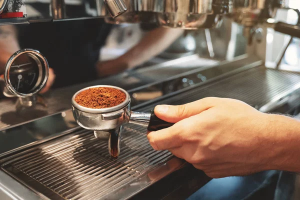 barista holding coffee holder with ground coffee near professional coffee machine preparing espresso in cafe close up