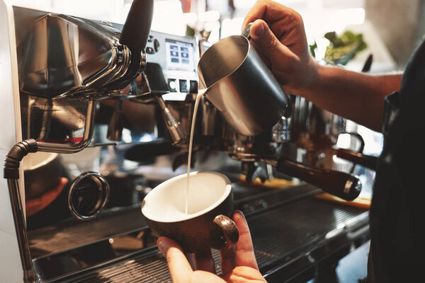 barista man pouring whipped milk from frothing pitcher in cup with coffee standing near professional coffee machine in cafe