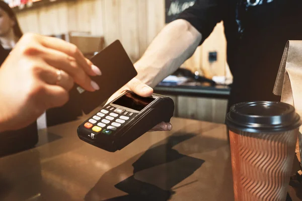 Barista hombre de pie detrás de la barra con bebida de café caliente en taza de papel y bolsa de papel de almuerzo para llevar la celebración de la terminal de pago para un cliente para comprar con tarjeta bancaria — Foto de Stock