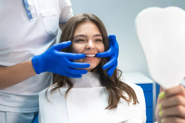 Hermosa joven morena mujer teniendo examen en el consultorio dental mientras dentista mostrando blanqueamiento resultados a paciente satisfecho — Foto de Stock