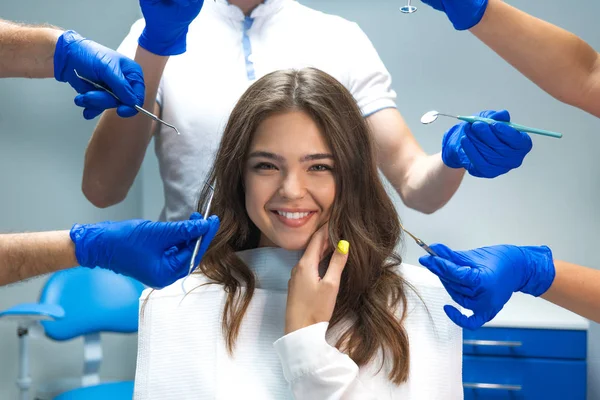 Feliz morena sonriente paciente sentada en silla de dentista rodeada de manos en guantes azules con instrumentos médicos y dentista en máscara de pie detrás — Foto de Stock